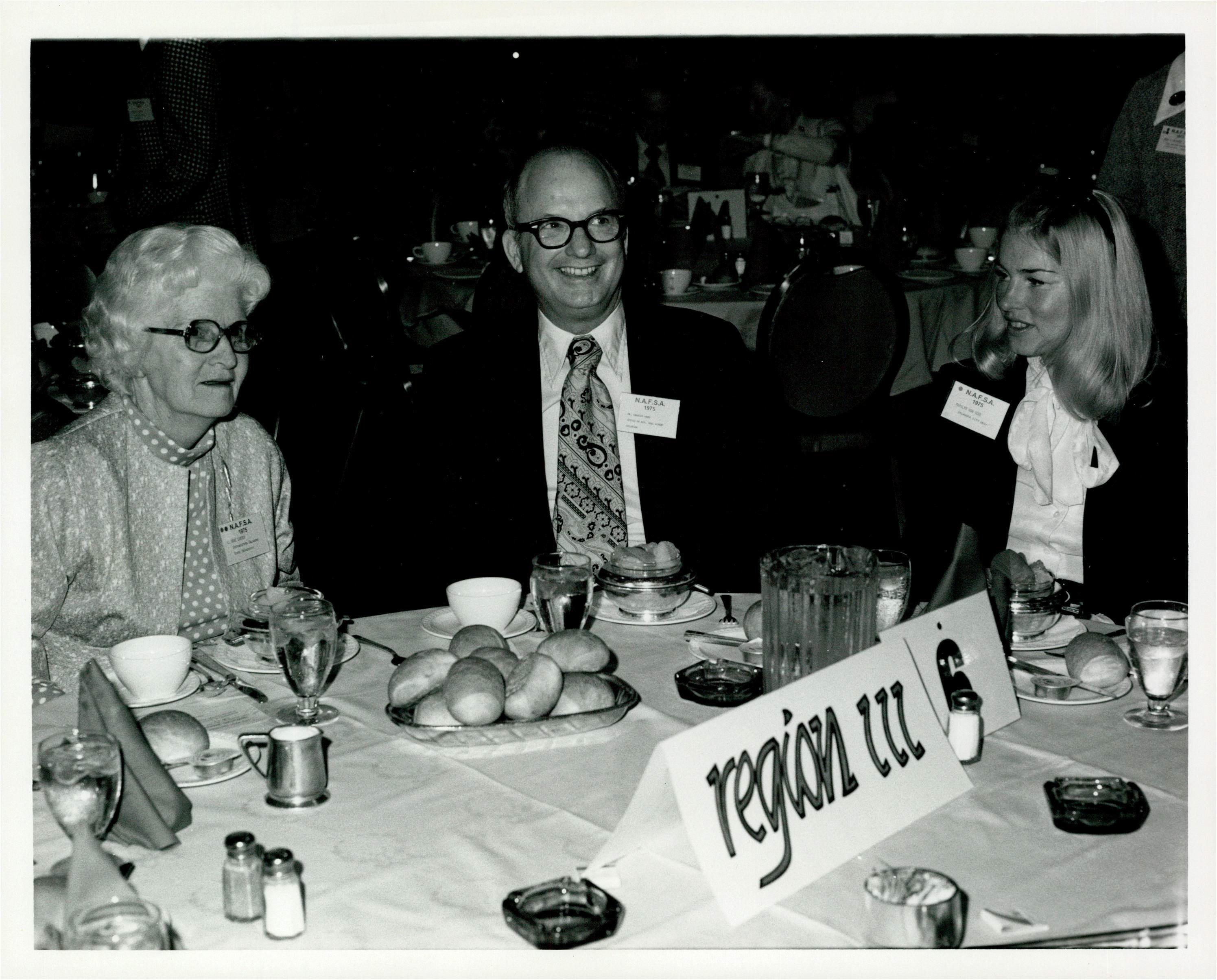 Black and white image of three people at a banquet sitting at a table with a sign that says region III 