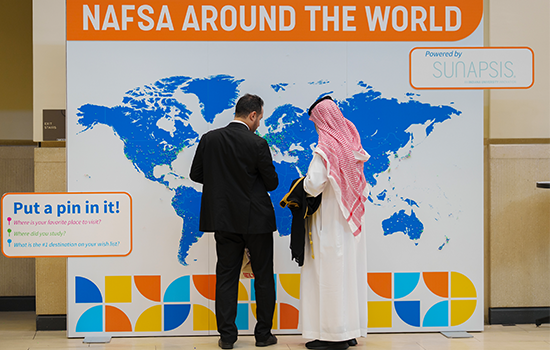 Two men, one in traditional middle eastern dress, stand in front of a map with pins for national origins