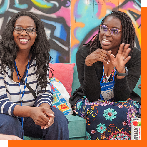 Two Black women sit talking in front of a colorful mural