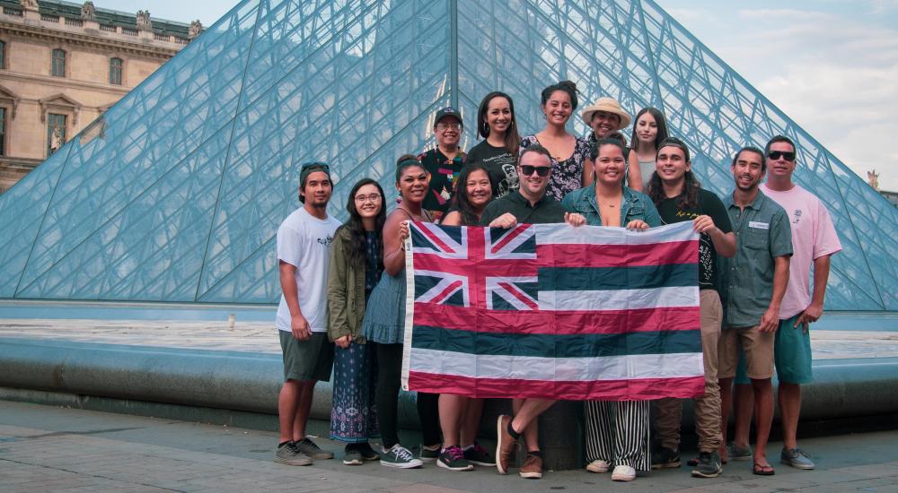 Students hold a Hawaiian flag in front of the Louvre museum.