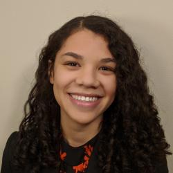 Caitlin Johnson headshot. Caitlin smiles to the camera in front of a beige background wearing a black blazer over a black and red blouse.