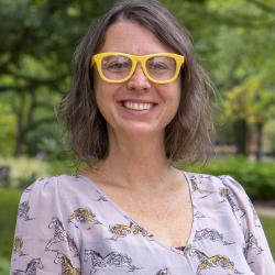 Cami Ross Headshot. Cami stands outside in a light purple top with yellow framed glasses. 