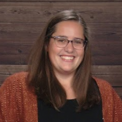 Headshot of Lindsey Goss smiling to the camera. Lindsey is has straight, brown hair, wears glasses, and poses in front of a dark wood paneled background. 