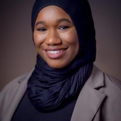 Malikka smiles in front of a gray background, wearing a black hijab and gray blazer