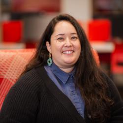 Phoebe sits in a red chair, smiling with a blue shirt on and green earrings 