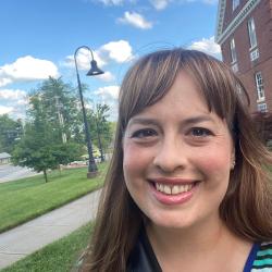 Katie smiles into the camera with a blue sky and green grass behind her. Katie has brown straight hair with bangs. 