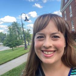 Katie smiles into the camera with a blue sky and green grass behind her. Katie has brown straight hair with bangs.