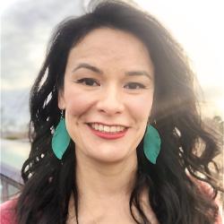 Woman with long black hair, pink top, and large green earrings, against sunny background.