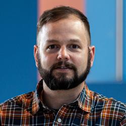 Man with beard and plaid shirt with a blue and orange striped wall in the background