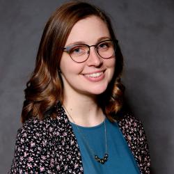 Natalie, with brown hair, glasses, a blue top, and floral-dot cardigan smiles to the camera in front of a grey backdrop