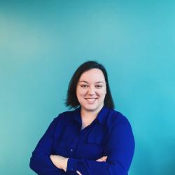 Sarah Warren smiles infront of a turqouise wall in a royal blue button up with arms crossed