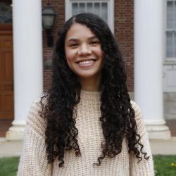 Caitlin wears a beige sweater, with long, dark curly hair, and white columns frame behind her.