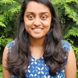 Kripa smiles in a blue dress in front of foliage