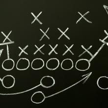 Photo of a person holding chalk drawing out a football play on a dark background