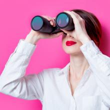 Woman looking through binoculars against a pink background